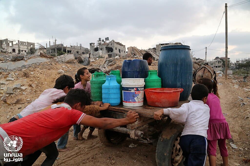 Children push a cart loaded with water containers past destroyed buildings in Gaza